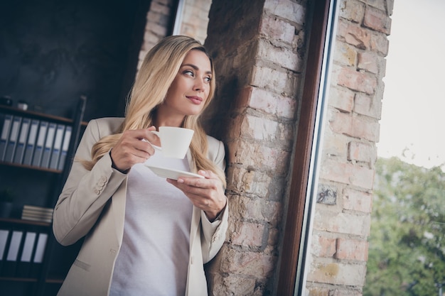 Cheerful business woman à loin de la fenêtre pour se détendre les yeux tout en se réchauffant avec une boisson chaude dans la tasse