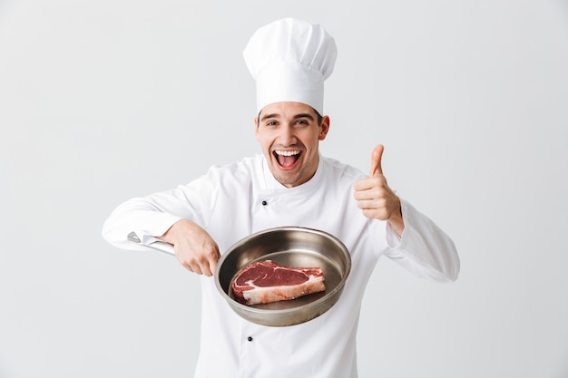 Cheerful chef cuisinier en uniforme montrant poêle avec un steak de boeuf cru isolé sur mur blanc, donnant les pouces vers le haut