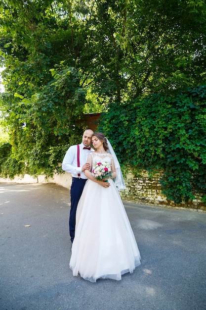 Cheerful couple bride dans une robe blanche avec un bouquet tandis que le marié avec bretelles et noeud papillon. Dans le contexte d'un mur avec des feuilles vertes. Couple heureux. Le concept de mariage.