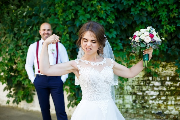 Cheerful couple bride dans une robe blanche avec un bouquet tandis que le marié avec bretelles et noeud papillon se tenant la main. Dans le contexte d'un mur avec des feuilles vertes. Couple heureux. Notion de mariage.