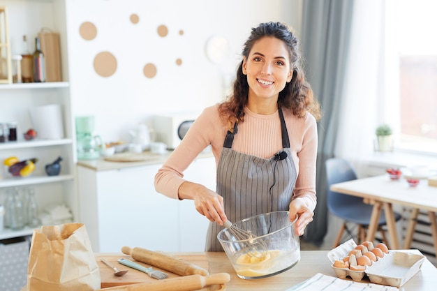 Cheerful Food Blogger Portant Un Tablier Debout à Table Dans Une Cuisine Moderne En Battant Des œufs Dans Un Bol En Verre à L'aide De Fouet, à La Recherche De Portrait