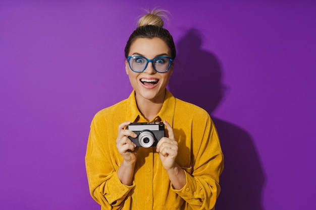 Cheerful girl wearing eyeglasses holding vintage film camera sur fond violet