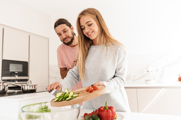 Cheerful joli couple cuisinant des repas sains ensemble tout en se tenant à la cuisine, tenant une planche à découper avec des légumes tranchés