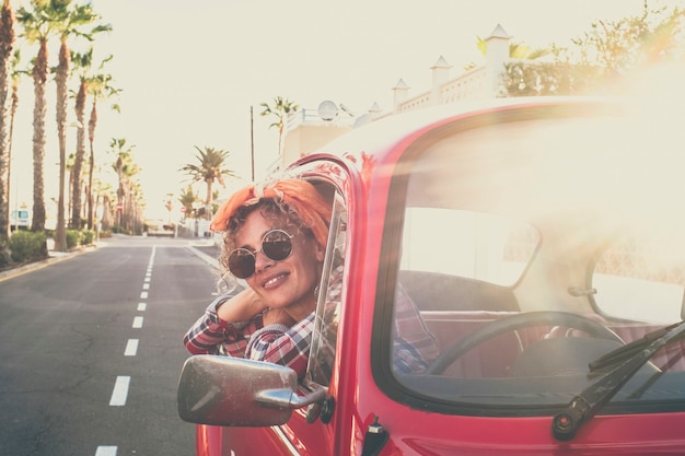 CheerfulÃ‚Â jolie jeune femme caucasienne adulte sourit et aime voyager debout à l'extérieur du vieux véhicule de voiture rouge à la recherche - des femmes à la mode avec des lunettes de soleil et des vêtements colorés - route en arrière-plan