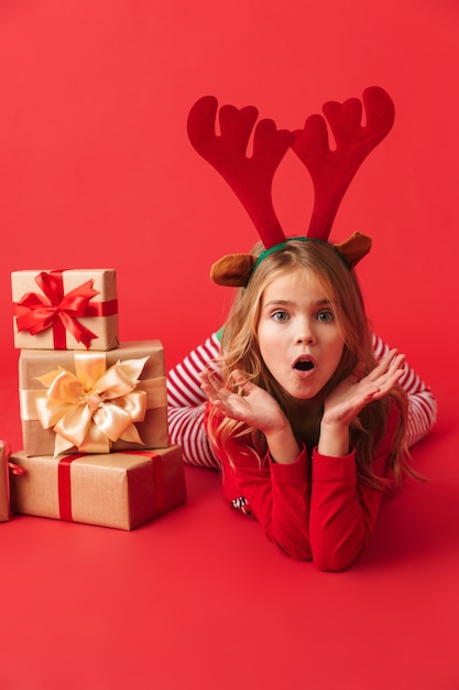 Cheerful little girl wearing Christmas raindeer costume assis avec pile de coffrets cadeaux isolés