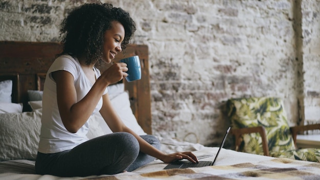 Cheerful mixed race girl tapant sur un ordinateur portable pour partager les médias sociaux assis dans son lit à la maison