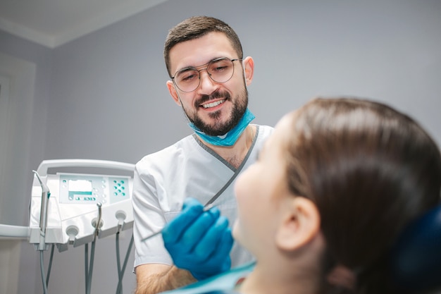 Cheerful nice male dentist tenir l'outil dans la main et sourire. Fille s'asseoir dans un fauteuil dentaire dans la chambre et regarder le médecin.