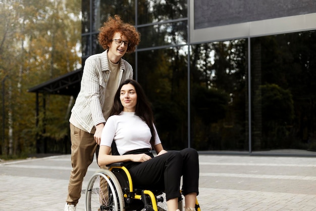 Cheerful redhaired curly guy in glasses and accolades porte un fauteuil roulant avec une fille Une belle brune est assise et regarde ailleurs dans la rue