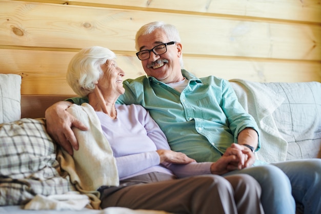 Cheerful senior couple in casualwear discuter de nouvelles ou de plans tout en vous relaxant sur un canapé dans leur maison de campagne