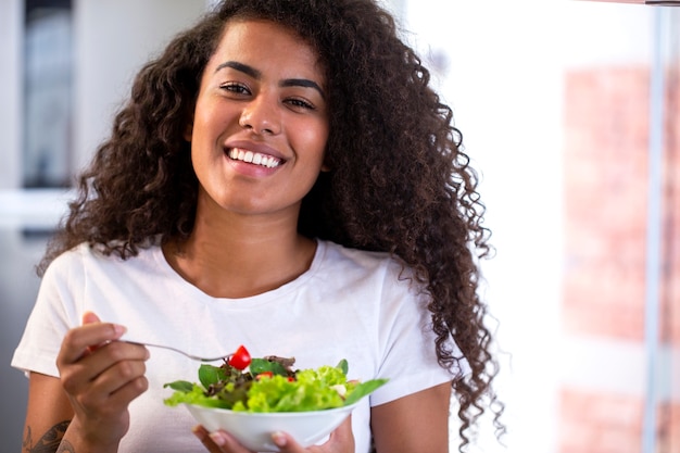 Cheerful young afro american woman eating salade de légumes dans la cuisine à domicile