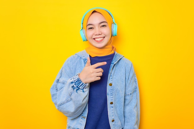 Cheerful young Asian woman in jeans jacket portant des écouteurs pour écouter de la musique isolé sur fond jaune