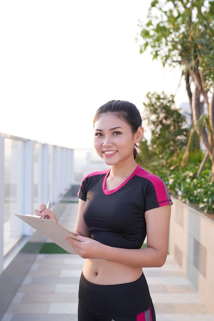 Cheerful young female fitness trainer écrit sur son presse-papiers en plein air.
