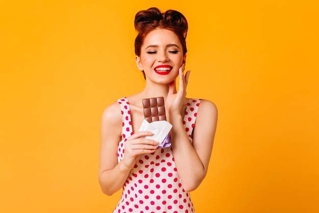 Cheerful young woman in polkadot dress holding chocolate Studio shot of happy pin-up souriant sur fond jaune