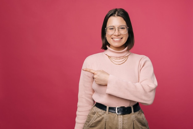 Cheerful young woman wearing glasses pointe de côté avec l'index sur fond rose Studio shot