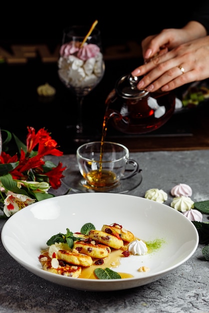 Cheesecake, confiture fraîche et menthe. Des crêpes au fromage cottage ou des beignets de caillé décorés de sucre en poudre en vue rapprochée de la plaque. Dans le contexte, la jeune fille verse du thé.