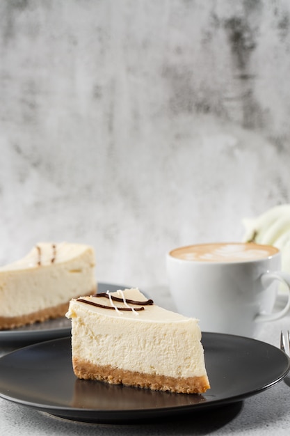 Cheesecake New York ou cheesecake classique avec une tasse de café sur le tableau blanc. Vue rapprochée. Petit déjeuner savoureux. Morceau de gâteau sur plaque noire, tasse blanche sur fond de marbre blanc. Photo verticale.