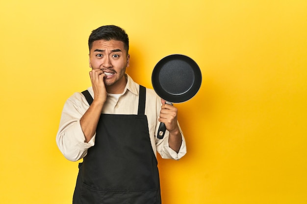 Un chef asiatique tenant une casserole en fond de studio jaune mordant les ongles nerveux et très anxieux