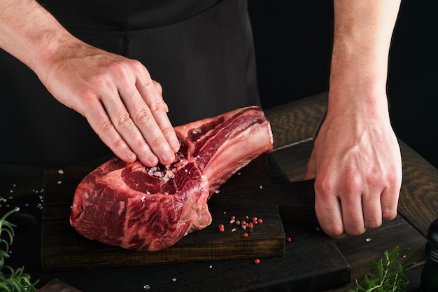 Chef coupant le steak de boeuf Les mains de l'homme tiennent le steak cru Tomahawk sur une planche à découper en bois rustique sur fond noir Recettes de cuisine et concept de restauration Mise au point sélective