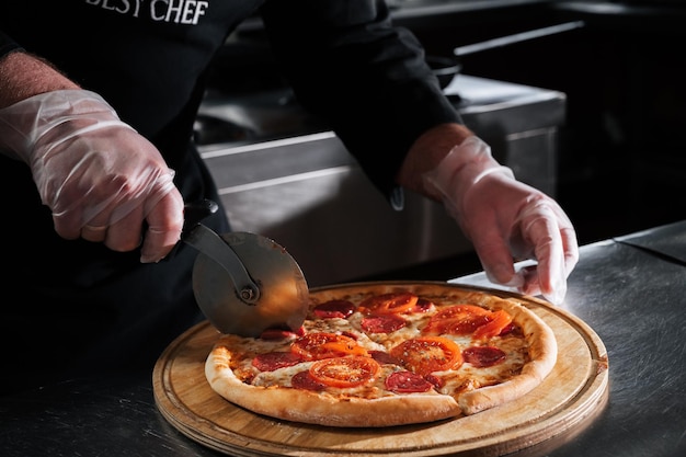Le chef coupe la pizza traditionnelle italienne au pepperoni en tranches avec un couteau dans la cuisine.