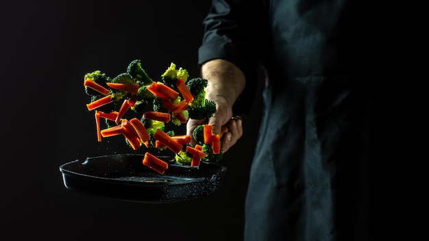 Chef cuisinant des légumes sur une casserole volant des légumes se dispersant dans un mouvement de gel d'un nuage en l'air sur fond noir