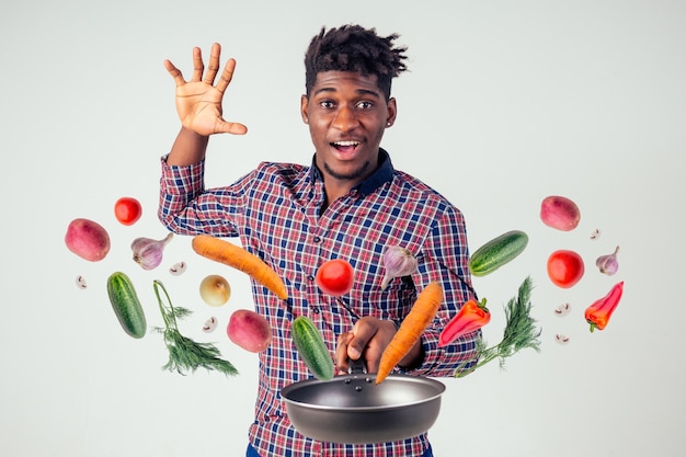 Chef cuisinier afro-américain tenant un homme sorcier de poêle à frire cuisinant une salade de nourriture volante magique, carotte, ail, oignon, poivre, pomme de terre, concombre isoler fond blanc studio. magie du goût fantaisie