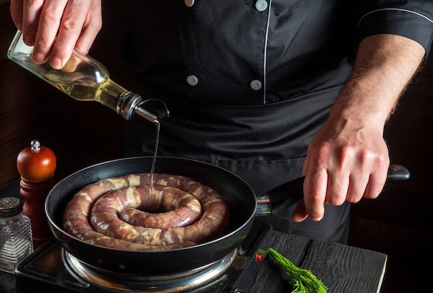 Le Chef Ou Le Cuisinier Ajoute De L'huile à La Poêle Avec Des Saucisses De Viande Crue. Préparation Pour La Cuisson Des Saucisses Dans La Cuisine D'un Restaurant Ou D'un Café Sur Une Table Avec Des Légumes