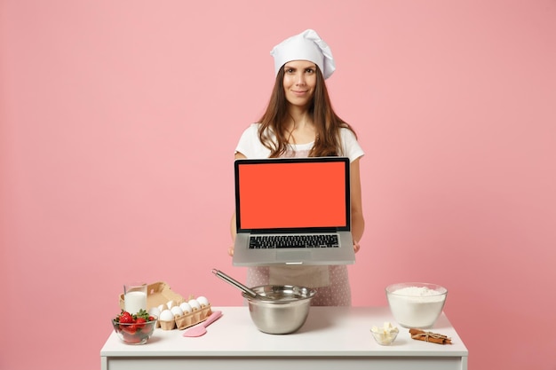 Chef cuisinier confiseur ou boulanger en t-shirt blanc, chapeau de chefs toque cuisine gâteau de Noël ou cupcake à table tenir pc ordinateur portable avec écran vide vide isolé sur fond rose pastel. Concept de maquette.