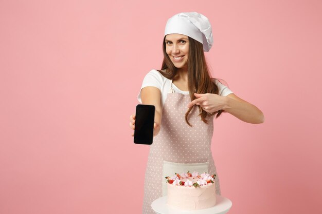 Chef cuisinier confiseur ou boulanger en t-shirt blanc tablier, chapeau de chefs toque cuisson gâteau ou cupcake à table tenir téléphone portable isolé sur fond pastel rose en studio. Maquette du concept de nourriture de l'espace de copie.