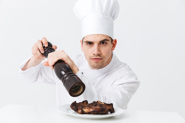 Chef cuisinier homme sérieux portant des poivrons uniformes steak de boeuf cuit sur une plaque isolée sur mur blanc