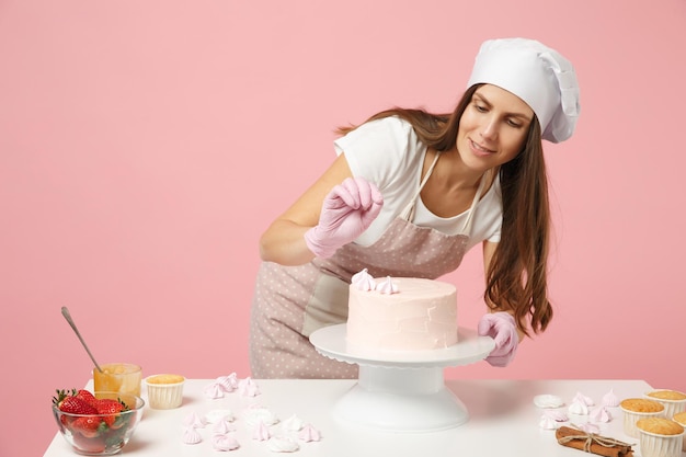 Chef cuisinier pâtissier boulanger en tablier blanc t-shirt toque chapeau de chefs cuisiner à table isolé sur fond pastel rose en studio. Processus de décoration de gâteau fraise meringuée. Maquette de concept alimentaire.