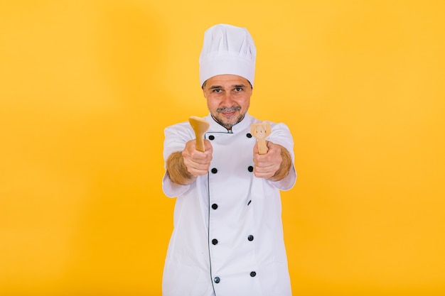 Chef cuisinier portant un bonnet de cuisine blanc et une veste avec cuillère et fourchette en bois, sur fond jaune