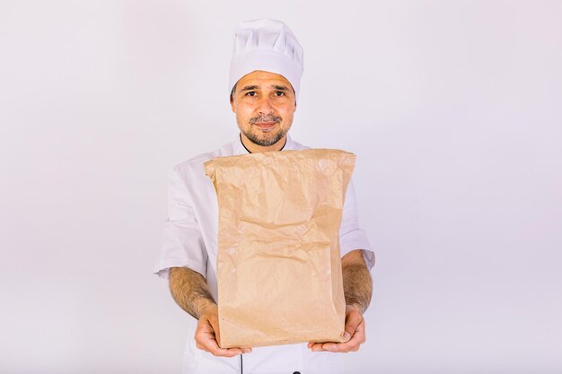 Chef cuisinier portant une casquette et une veste de cuisine blanches, tenant un sac en papier avec des plats à emporter, sur fond blanc