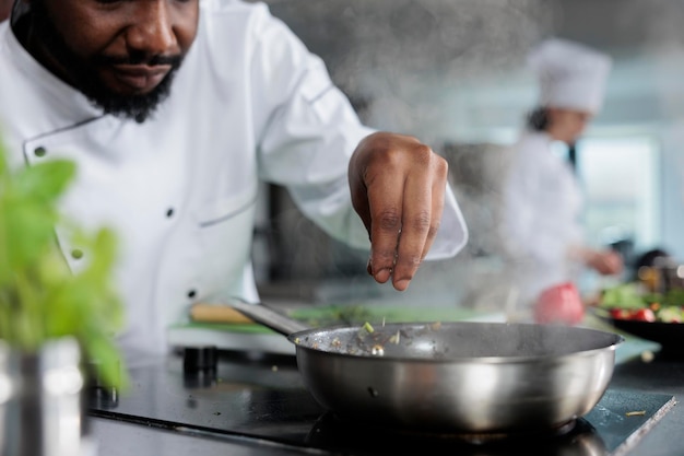 Chef cuisinier portant un uniforme de cuisine garnissant un plat gastronomique avec des herbes fraîches hachées dans la cuisine du restaurant. Expert en gastronomie utilisant une poêle pour cuisiner de délicieux plats pour le service du dîner.