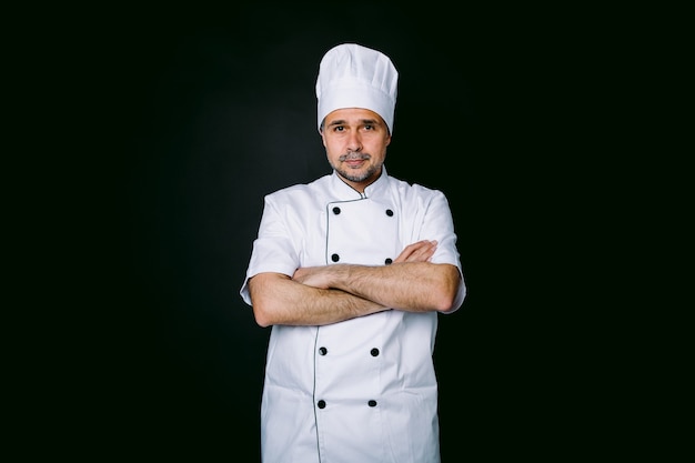 Chef cuisinier portant une veste et un chapeau de cuisine, les bras croisés, sur fond noir