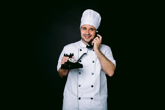 Chef cuisinier portant une veste et un chapeau de cuisine, parlant sur un téléphone rétro, sur fond noir