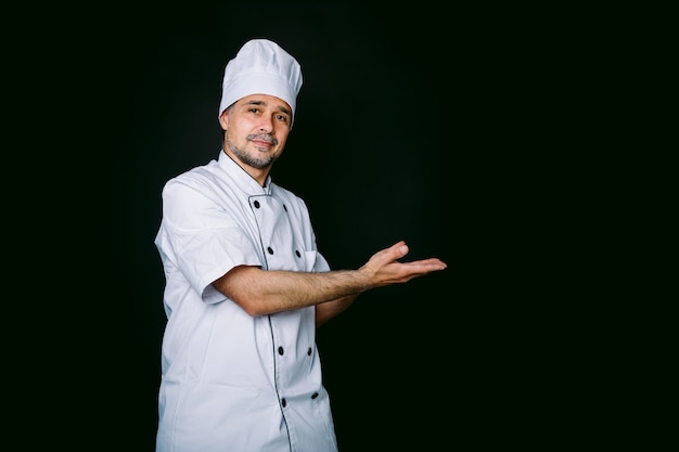 Chef cuisinier portant veste et chapeau de cuisine pointant vers la droite sur fond noir