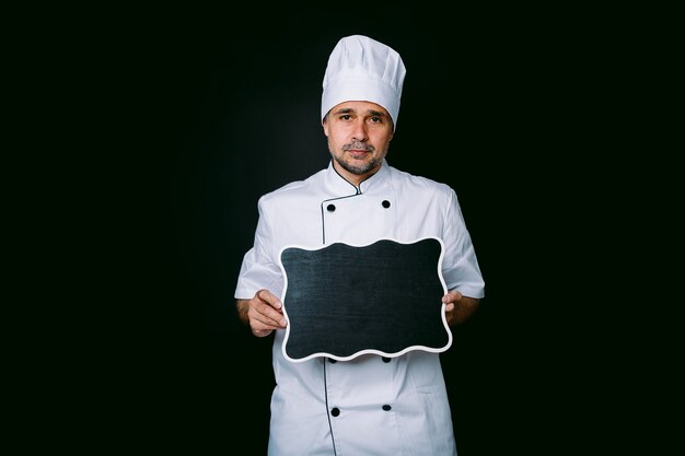 Photo chef cuisinier portant une veste et un chapeau de cuisine, tenant un tableau noir avec menu, sur fond noir