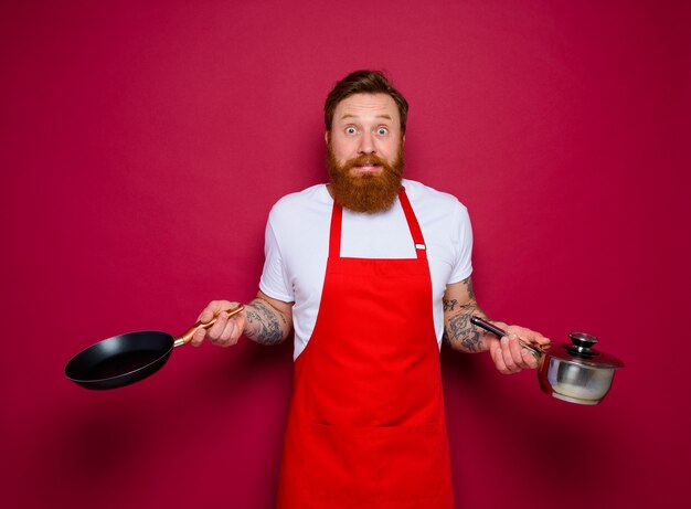 Le chef effrayé avec la barbe et le tablier rouge cuisine avec la casserole et le pot