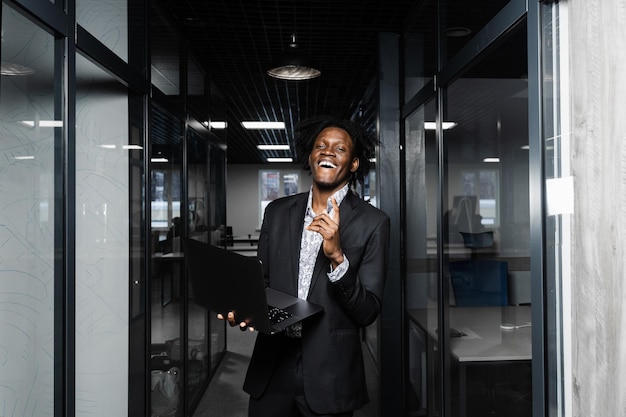 Un chef d'entreprise noir joyeux avec un ordinateur portable pointe les doigts vers le haut et sourit au bureau.