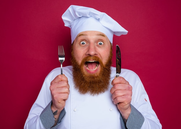 Le chef étonné avec la barbe et le tablier rouge tient des couverts à disposition