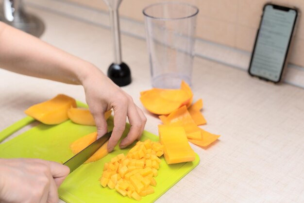 Chef féminin coupant de la mangue fraîche pour faire de délicieux smoothies ou jus de mangue faits maison à l'aide d'une recette en ligne à partir d'un smartphone Mise au point sélective Processus de fabrication d'une boisson d'été à la maison mode de vie sain