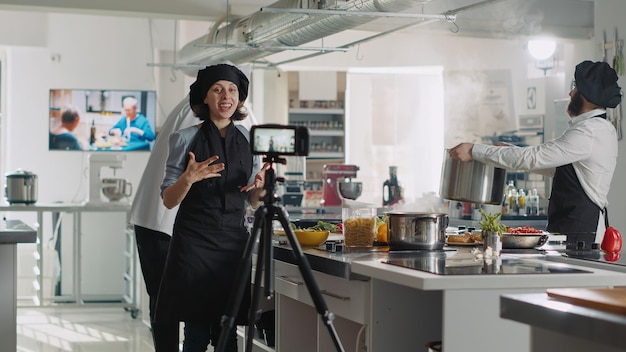 Chef féminin enregistrant une vidéo d'émission de cuisine sur un programme télévisé, filmant un vlog culinaire avec caméra dans la cuisine d'un restaurant professionnel. Femme en uniforme parlant de gastronomie lors d'un cours de cuisine en ligne.