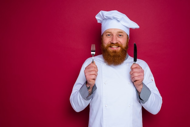 Le chef heureux avec la barbe et le tablier rouge tient des couverts à disposition