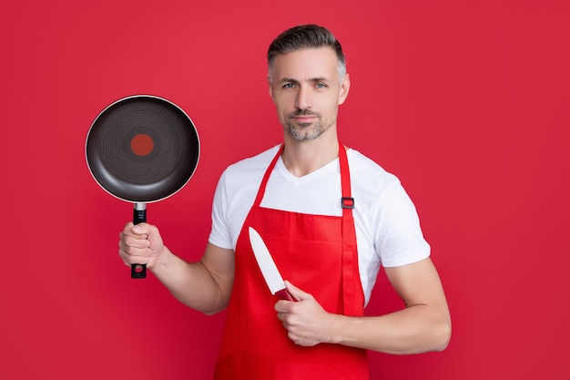 Chef d'homme mûr en tablier avec poêle et couteau sur fond rouge