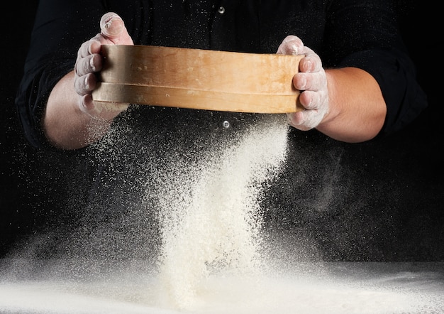 Chef un homme en uniforme noir tient un tamis en bois rond dans ses mains et tamise la farine de blé blanc sur fond noir