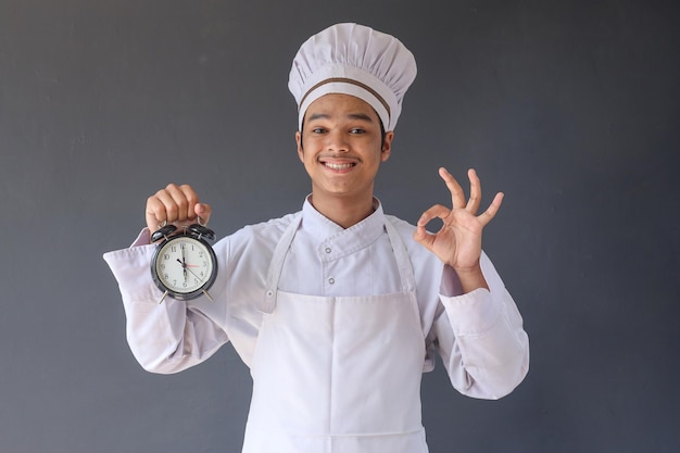 Chef masculin asiatique ou homme boulanger en chemise uniforme blanche tenant l'horloge et montrant le signe correct isolé sur