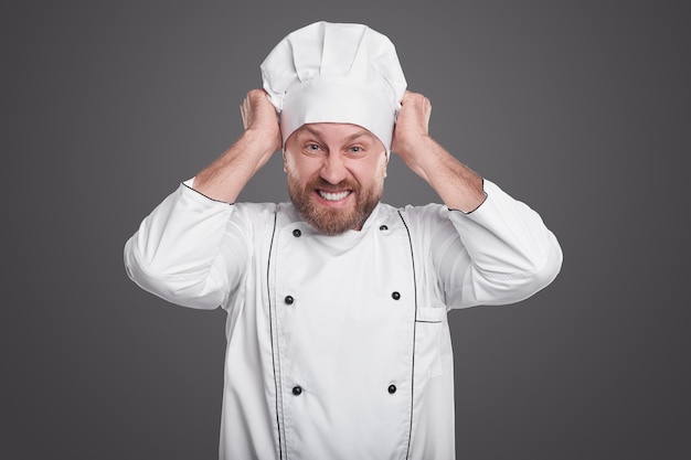 Chef masculin barbu furieux regardant la caméra et tirant le chapeau dans la colère tout en travaillant au restaurant sur fond gris