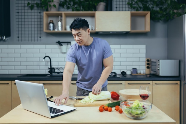 Le chef masculin cuisine à la maison et enseigne en ligne des légumes asiatiques coupés dans la cuisine utilise un ordinateur portable pour des cours de cuisine en ligne