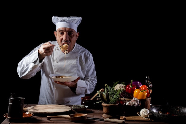 Chef masculin dans une cuisine de restaurant vêtu d'un uniforme blanc dégustant des spaghettis avec des légumes frais avant de le servir