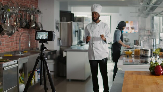 Chef masculin enregistrant une émission de cuisine vlog télévisé sur caméra, parlant de gastronomie et de recette culinaire. Jeune cuisinier filmant du contenu de cuisine professionnelle pour un programme de télévision en ligne. Prise de vue au trépied.